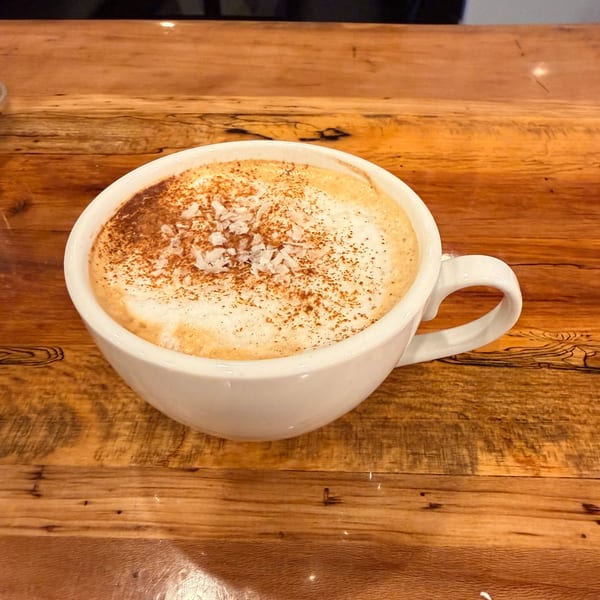 A white mug of cappuccino with foam, cinnamon, and coconut flakes on a rustic wooden table.