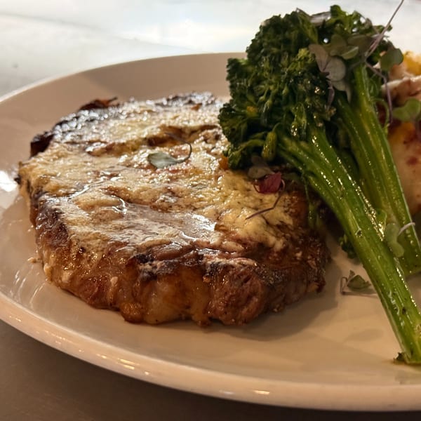 Grilled steak topped with melted cheese, served with vibrant green broccolini on a white plate.
