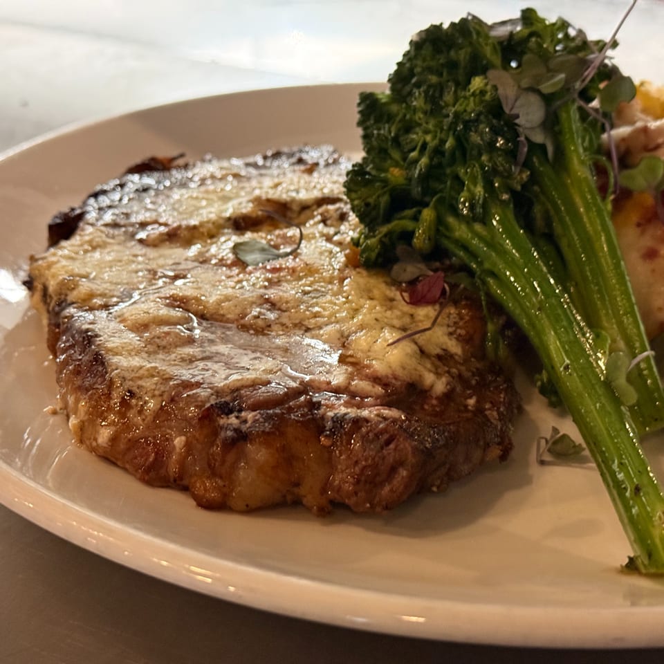 Grilled steak topped with melted cheese, served with vibrant green broccolini on a white plate.