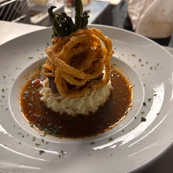 A Live Fire Chefs dish with mashed potatoes, gravy, fried onions, and asparagus on a white plate.